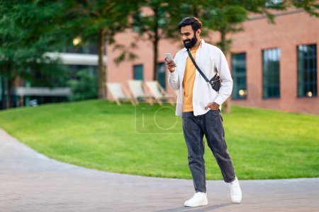 Photo for Full length of arabic guy with smartphone gadget, browsing internet and texting while walking outdoor in urban area in evening, using mobile application via phone for communication. Copy space - Royalty Free Image