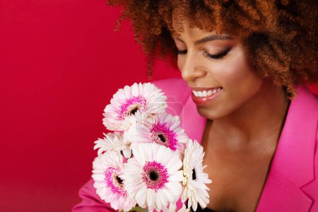 Foto de Estudio Retrato de la atractiva joven negra posando con flores sobre fondo rojo. Primer plano de Bushy mujer afroamericana sosteniendo Gerberas flores cerca de la cara. Concepto de belleza femenina - Imagen libre de derechos