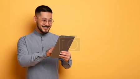 Smiling Young Asian Man Using Digital Tablet While Standing Over Yellow Studio Background, Handsome Millennial Man In Eyeglasses Browsing New App Or Shopping Online On Modern Gadget, Copy Space