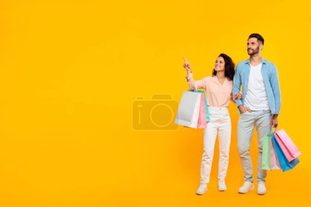 Seasonal discounts. Happy spouses standing with shopping bags and pointing aside at free space, posing on yellow background, european couple enjoying shopper deals, copy space