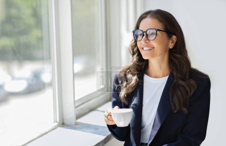 Photo for Successful cheerful attractive mature businesswoman in formal outfit wearing eyeglasses standing next to window with mug in her hand, drinking coffee at office, enjoying view, copy space - Royalty Free Image