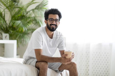 Foto de Feliz joven indio que se relaja en la cama con una taza de café, Sonriendo Millennial Oriental Guy Descansando en el acogedor dormitorio en casa, Hombre alegre disfrutando de la bebida caliente, Primer plano Shot con espacio de copia - Imagen libre de derechos