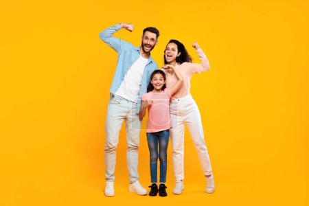 Foto de Fuerza familiar. feliz europeo padres y hija mostrando bíceps músculos y sonriendo a la cámara, posando juntos de pie sobre amarillo estudio fondo, longitud completa - Imagen libre de derechos