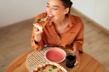 Foto de Mujer emocionada disfrutando de comer pizza sabrosa, sosteniendo y mordiendo rebanada, sentado a la mesa en el interior de la cocina, espacio libre, por encima de la vista. Comida de engaño, nutrición y comida rápida - Imagen libre de derechos