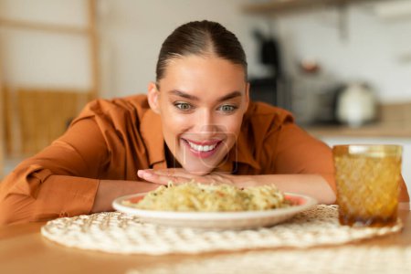 Foto de Jovencita alegre sentada en la mesa mirando deliciosa pasta en el plato disfrutando de la comida entregada, emocionada señora hambrienta quiere comer espaguetis, interior de la cocina - Imagen libre de derechos