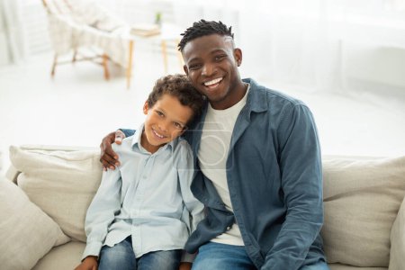 Foto de Retrato de feliz joven negro papá y pequeño hijo abrazando posando juntos en el interior de la sala de estar moderna, papá abrazando a su niño y sonriendo a la cámara, sentado en el sofá en casa - Imagen libre de derechos