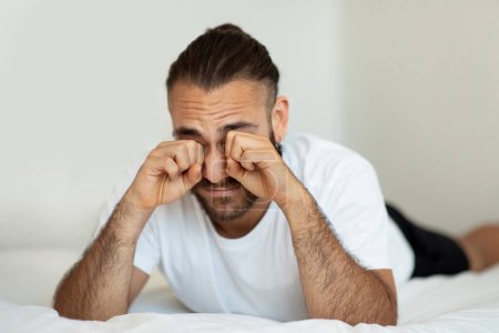 Photo for Young bearded man waking up in bed, rubbing eyes after sleepless night, stressed guy suffering from insomnia or headache while lying in bedroom at home, closeup shot with free space - Royalty Free Image