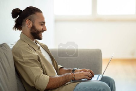 Téléchargez les photos : Vue latérale de joyeux beau barbu aux cheveux longs assis sur le canapé à la maison, tapant sur le clavier de l'ordinateur portable et souriant, espace de copie. Homme heureux entrepreneur indépendant développeur travaillant à distance - en image libre de droit