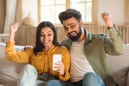 Téléchargez les photos : Couple indien joyeux utilisant smartphone et secouer les poings, lire de bonnes nouvelles, célébrer la grande chance et sourire, assis sur le canapé à la maison. Grande offre mobile, application pour téléphone portable concept de publicité - en image libre de droit