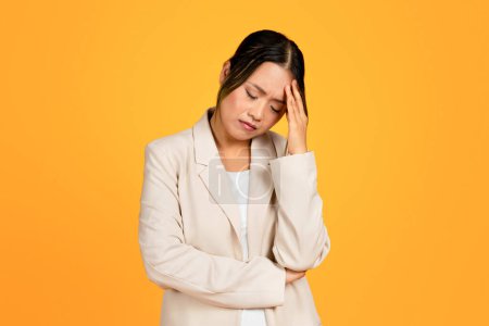 Foto de Mujer asiática joven triste en traje presiona mano a cabeza, que sufre de dolor de cabeza, aislado en el fondo del estudio amarillo. Migraña, problemas de salud, emociones, estrés, presión - Imagen libre de derechos