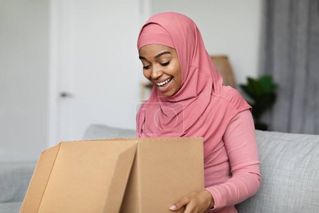 Téléchargez les photos : Satisfait femme musulmane noire ouverture boîte en carton, excité par un excellent service de livraison, assis sur le canapé à la maison, espace de copie. Jeune femme en hijab déballer colis au salon - en image libre de droit