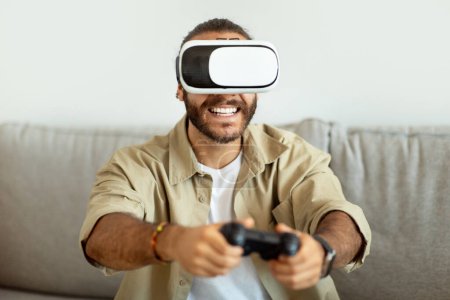 Téléchargez les photos : Divertissement, jeux, loisirs. Jeune homme barbu souriant portant une tenue décontractée assis sur le canapé, utilisant des lunettes VR et tenant le joystick de la console, jouant à des jeux vidéo à la maison, copiant l'espace - en image libre de droit