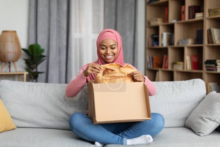 Foto de Compra rápida en tienda online. Señora musulmana negra satisfecha abriendo la caja de cartón, cliente feliz tomando la ropa de paquete, sentado en el sofá en la sala de estar en casa, espacio de copia - Imagen libre de derechos