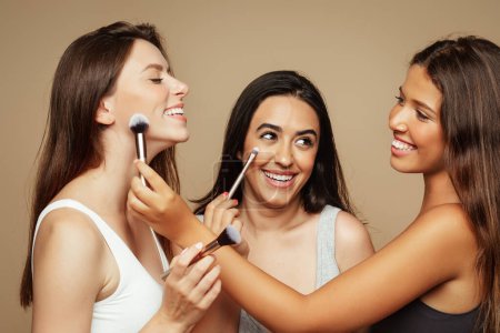Photo for Cheerful millennial latin woman in underwear apply nude makeup with brushes to caucasian lady, enjoy beauty care, isolated on beige background, studio. Cosmetics, lesson and help - Royalty Free Image