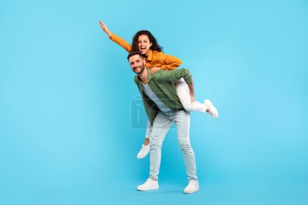 Photo for Handsome man giving piggyback ride to his wife against blue studio background, full length portrait, free space. Excited couple having fun, spending romantic times together - Royalty Free Image