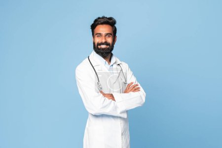 Téléchargez les photos : Portrait d'un Indien souriant d'âge moyen, homme médecin posant les mains croisées sur fond bleu studio, souriant à la caméra. Médecin généraliste professionnel - en image libre de droit