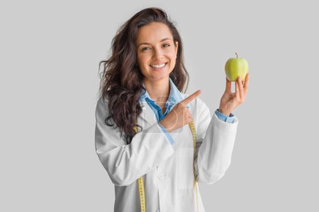 Fröhliche junge Ärztin in Uniform zeigt mit dem Finger auf einen Apfel auf hellgrauem Hintergrund und lächelt in die Kamera. Gesundheit, Ernährung, Ernährung, Beratung durch Ernährungsberater