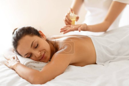 Foto de Tratamiento de Relajación Spa. Relajado Millennial Lady disfrutando del masaje con aceites en el centro de spa, Masajista mujer hidratante cuerpo y piel aplicación de aceite para el cuidado de la piel, enfoque selectivo - Imagen libre de derechos