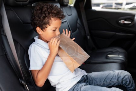 Photo for Sick black boy in the backseat of a car sitting alone covers his mouth, breathing in paper bag. School african american guy kid suffers from motion sickness during road trip, side view - Royalty Free Image