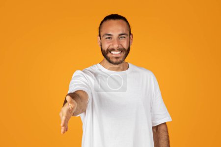 Photo for Smiling friendly adult european guy with beard in white t-shirt gives hand for shake, isolated on orange studio background. Say hello, introduce yourself, meeting and welcome, lifestyle - Royalty Free Image