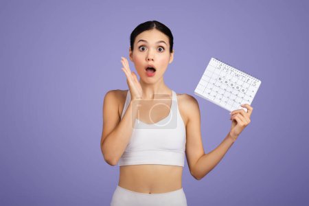Photo for Shocked young caucasian lady in sportswear with open mouth showing calendar, posing on purple studio background. Sports and menstruation, weight loss and problems with workout - Royalty Free Image