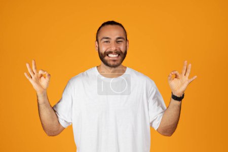 Foto de Sonriente chico europeo adulto con barba en camiseta blanca haciendo signo de ok con las manos, aislado sobre fondo de estudio naranja. Recomendación, consejo, venta y estilo de vida, emociones humanas, anuncio y oferta - Imagen libre de derechos