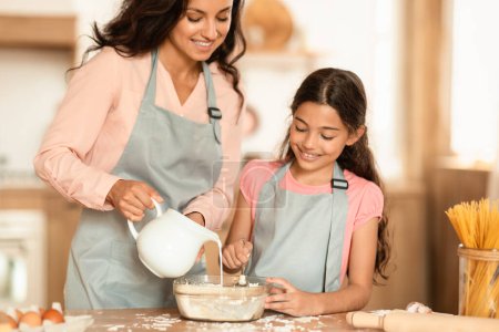 Photo for Smiling Mom And Preteen Daughter Baking Making Dough For Cookies, Adding Milk In Bowl Standing In Home Kitchen, Wearing Aprons. Family Pastry Recipes Concept. Cropped Shot - Royalty Free Image