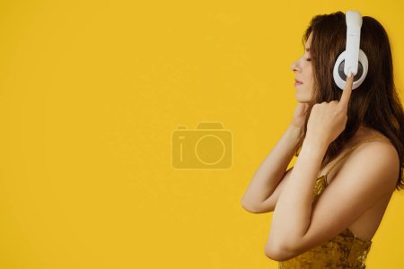 Téléchargez les photos : Femme calme européenne écoutant de la musique dans des écouteurs sans fil avec les yeux fermés et un casque tactile, profitant de chansons préférées tout en se tenant sur fond jaune, vue latérale, espace libre - en image libre de droit