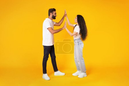 Foto de Longitud completa de amantes cónyuges árabes aplaudiendo y sosteniendo las manos, celebrando el logro y el éxito, dando High Five con expresión gozosa sobre fondo amarillo, Studio Shot - Imagen libre de derechos