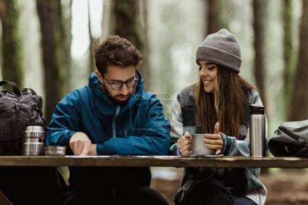 Photo for Happy young caucasian lady and man in jackets drinking coffee, planning route with map, enjoy adventure in cold forest in autumn. Spare time, hikers vacation together, active lifestyle and rest - Royalty Free Image
