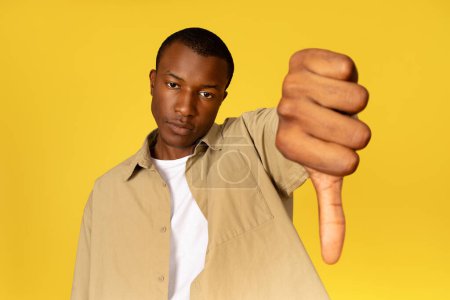 Photo for Sad offended dissatisfied young african american guy in casual wear showing thumbs down isolated on yellow studio background. bad review, service problems, advertising and offer - Royalty Free Image