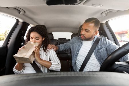 Photo for Sick black lady sitting in the front seat of a car covers mouth, breathing in paper bag. Dizzy african american woman suffers from motion sickness during road trip, concerned man husband comfort her - Royalty Free Image