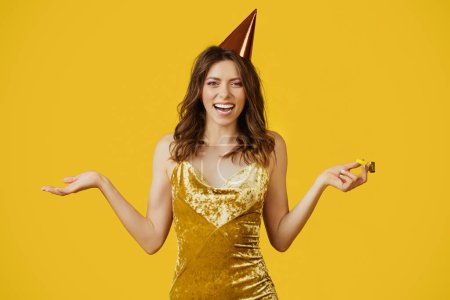 Photo for Satisfied young woman in elegant dress and birthday cap holding party horn, looking and smiling at camera, celebrating her special day on yellow studio background - Royalty Free Image