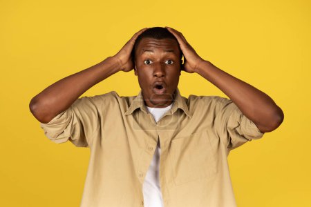 Photo for Shocked frightened young african american guy in casual presses hands to head, isolated on yellow studio background. Fear, horror, stress and bad news reaction, ad and offer - Royalty Free Image