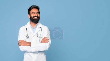 Photo for Confident cheerful indian male doctor in white uniform posing with folded arms and looking aside at free space over blue studio background, panorama. Physician with stethoscope around neck - Royalty Free Image