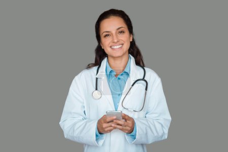 Téléchargez les photos : Docteur femme tenant smartphone à l'aide d'une application médicale, debout sur fond de studio gris, souriant à la caméra. Rendez-vous à distance avec un médecin professionnel concept - en image libre de droit