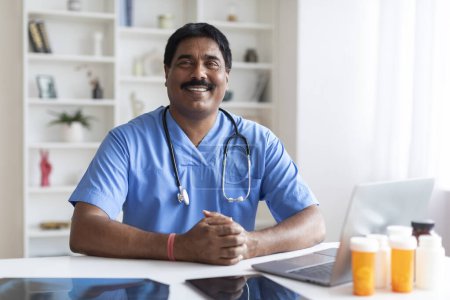 Foto de Médico masculino indio sonriente en uniforme azul sentado en el escritorio con el ordenador portátil en el lugar de trabajo, hombre terapeuta profesional que usa exfoliantes médicos y estetoscopio posando en la oficina en la clínica moderna, espacio de copia - Imagen libre de derechos