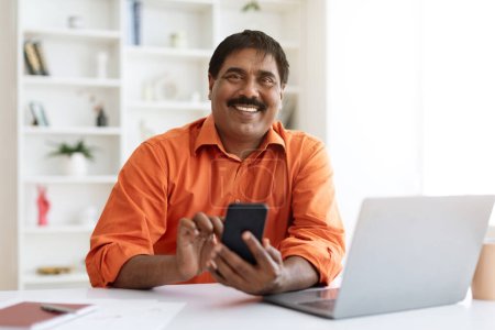 Photo for Positive mature indian man in smart casual sitting at workplace, using computer laptop and smartphone at office, using business mobile application. Hindu entrepreneur working from home, copy space - Royalty Free Image