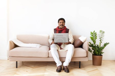 Téléchargez les photos : Heureux homme indien mature en costume national entrepreneur indépendant assis sur le canapé, en utilisant un ordinateur portable à la maison. Homme d'affaires de l'Est commerçant sur les marchés boursiers en ligne, travaillant à distance, l'espace de copie - en image libre de droit