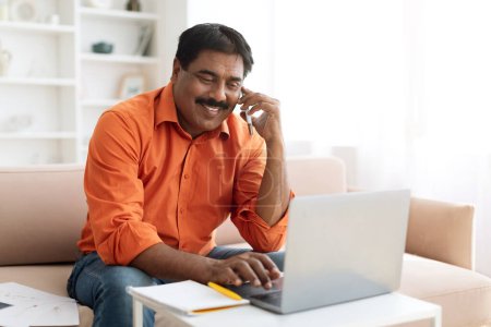 Téléchargez les photos : Homme d'affaires indien d'âge moyen portant une tenue décontractée travaillant à la maison, assis sur le canapé dans le salon, utilisant un ordinateur portable, avoir une conversation téléphonique avec le client, partenaire d'affaires, espace de copie - en image libre de droit