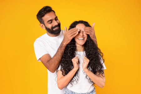 Photo for Romantic surprise. Cheerful middle eastern spouses posing, man covering wifes eyes surprising standing behind her back over yellow studio background. Anniversary congratulations - Royalty Free Image