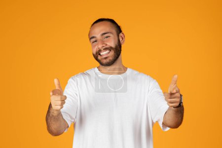 Téléchargez les photos : Heureux homme européen adulte confiant avec barbe en t-shirt blanc doigt pointeur à la caméra, isolé sur fond de studio orange. Motivation, conseils professionnels, votre choix, votre tour et mode de vie - en image libre de droit