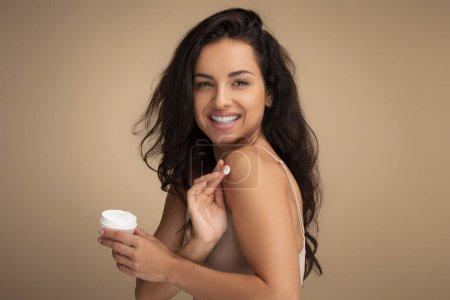 Photo for Happy beautiful brunette young indian woman wearing beige top applying body cream or lotion on shoulder and smiling at camera, isolated on color background. Skin moisturizing, body care concept - Royalty Free Image