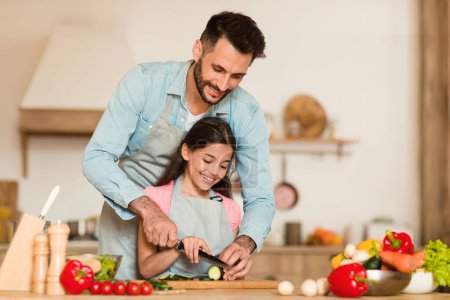 Foto de Cocinar con papá. Chica feliz preparando la cena con su padre en la cocina, amando a papá haciendo ensalada de verduras con su niña preadolescente, espacio libre - Imagen libre de derechos