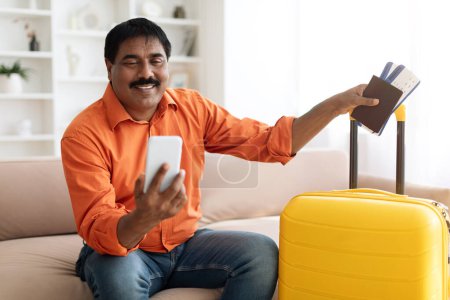 Photo for Excited mature indian man tourist check-in by application on phone from home, sitting on couch in living room, holding yellow suitcase, passport and tickets, going vacation. Travelling mobile app - Royalty Free Image
