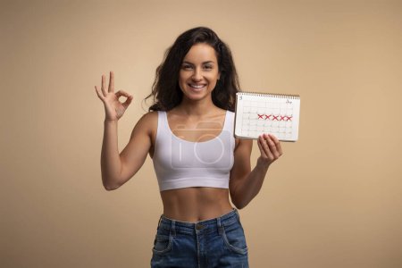 Photo for Happy cheerful young brunette woman demonstrates periods calendar and showing okay gesture, isolated on beige studio background. Women healthcare, menstruation, menstrual cycle concept - Royalty Free Image