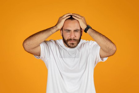 Photo for Sad upset disappointed adult european guy in white t-shirt presses hands to head, suffers from migraine, find mistake, isolated on orange studio background. Headache, stress, pressure and emotions - Royalty Free Image