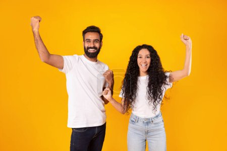 Photo for Victory Celebration. Happy Middle Eastern Spouses Shaking Fists With Joyful Expression, Raising Arms And Smiling To Camera Posing In Studio Over Yellow Background. Common Success, Achievement - Royalty Free Image