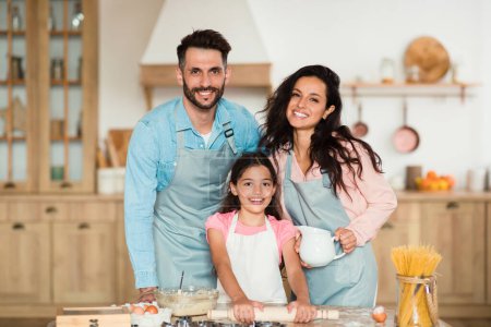 Téléchargez les photos : Portrait d'une heureuse famille européenne de trois personnes posant pendant la cuisson de pâtisseries dans la cuisine ensemble, mère, père et leur fille souriant à la caméra, espace de copie - en image libre de droit