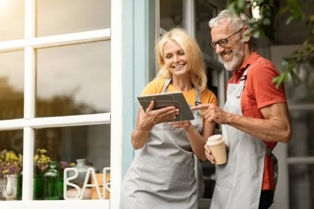Photo for Modern Technologies. Smiling Mature Farmers Couple In Aprons Using Digital Tablet Together And Drinking Coffee While Standing At Terrace Near Country House, Happy Spouses Browsing Internet On Gadget - Royalty Free Image